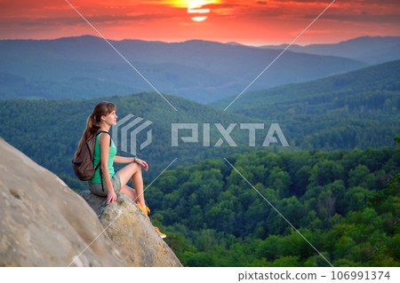 Tired hiker girl relaxing on rocky mountain top enjoying evening nature during travelling on wilderness trail. Lonely woman traveler traversing high hilltop route. Healthy lifestyle concept 106991374