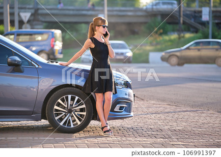 Stylish woman driver standing near her vehicle talking on cellphone on city street in summer Stylish woman driver standing near her vehicle talking on cellphone on city street in summer 106991397