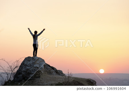 Silhouette of a woman hiker standing alone on big stone at sunset in mountains. Female tourist raising her hands up on high rock in evening nature. Tourism, traveling and healthy lifestyle concept. 106991604