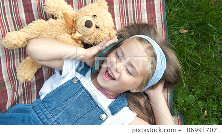 Pretty child girl laying on green lawn with her teddy bear talking on mobile phone. 106991707