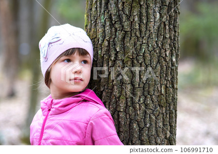 Portrait of young pretty child girl wearing pink jacket and cap leaning to a tree in forest enjoying warm sunny day in early spring outdoors. 106991710