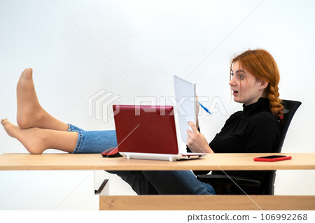 Happy young office worker woman sitting relaxed with feet on table behind working desk with laptop computer, cell phone and notebook. 106992268