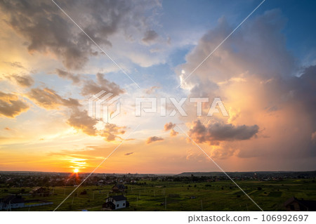 Dramatic sunset over rural area with stormy puffy clouds lit by orange setting sun and blue sky. 106992697