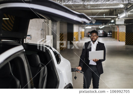 Man with computer plugging in EV charging cord in parking Man with computer plugging in EV charging cord in parking 106992705