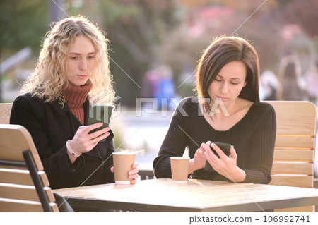 Distracted women browsing their sellphones sitting together at city street cafe. Girlfriends ignoring each other while looking in gadgets. Lack of personal contact between friends in real life concept Distracted women browsing their sellphones sitting together at city street cafe. Girlfriends ignoring each other while looking in gadgets. Lack of personal contact between friends in real life concept 106992741