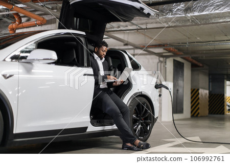 Businessman sitting in e-car on charge with laptop on knees 106992761