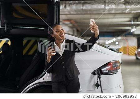 Lady with drink smiling at camera near charging EV in garage Lady with drink smiling at camera near charging EV in garage 106992958