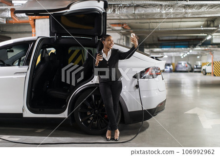 Woman with cup taking selfie on mobile during e-car charging Woman with cup taking selfie on mobile during e-car charging 106992962