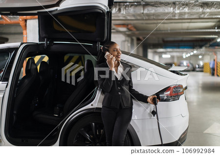 Lady chatting via gadget while charging e-car in parking Lady chatting via gadget while charging e-car in parking 106992984