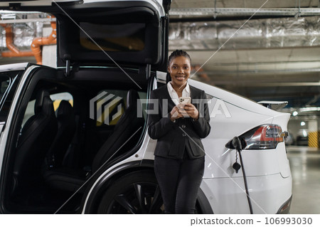 Businesswoman with mobile waiting for e-car being filled up Businesswoman with mobile waiting for e-car being filled up 106993030
