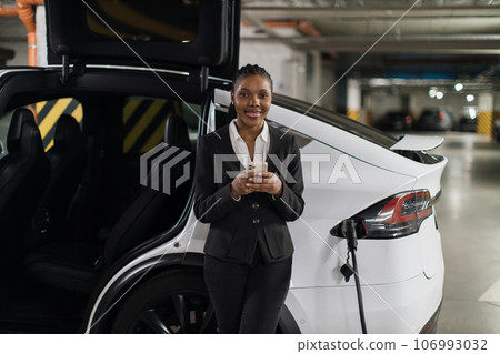 Businesswoman with mobile waiting for e-car being filled up Businesswoman with mobile waiting for e-car being filled up 106993032