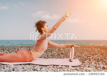 Woman sea pilates. Sporty happy middle aged woman practicing fitness on beach near sea, smiling active female training with ring on yoga mat outside, enjoying healthy lifestyle, harmony and meditation 106993169