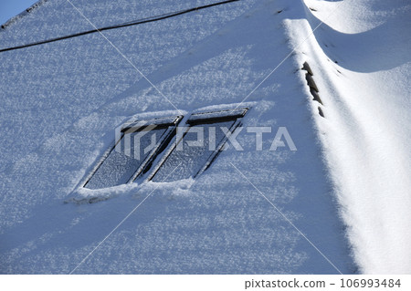 Closeup of house roof top with attic windows covered with snow in cold winter. Tiled covering of building in wintertime weather Closeup of house roof top with attic windows covered with snow in cold winter. Tiled covering of building in wintertime weather 106993484