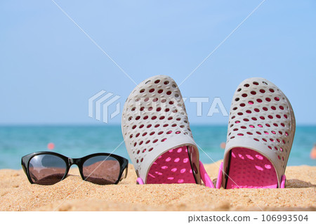 Closeup of clogs shoes and black protective sunglasses on sandy beach at tropical seaside on warm sunny day. Summer vacation concept Closeup of clogs shoes and black protective sunglasses on sandy beach at tropical seaside on warm sunny day. Summer vacation concept 106993504