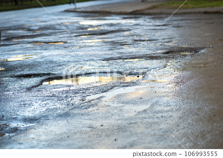 Close up of a road in very bad condition with big potholes full of dirty rain water pools. 106993555