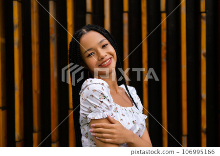 Portrait of a young pretty afro woman with pigtails who smiles, looks at the camera and enjoys herself, posing half sideways and hugging her hand Portrait of a young pretty afro woman with pigtails who smiles, looks at the camera and enjoys herself, posing half sideways and hugging her hand 106995166