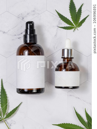 Cosmetic bottles with blank labels near green cannabis leaves on marble table. Mockup 106996891