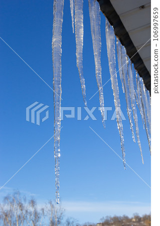 Icicles hang on the roof of a house against a bright blue sky. Spring landscape with icicles hanging from the roof of the house. Set of snow icicles, snow hat Icicles hang on the roof of a house against a bright blue sky. Spring landscape with icicles hanging from the roof of the house. Set of snow icicles, snow hat 106997659