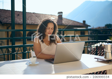 Confident female real estate agent working on laptop from home balcony, against a beautiful landscape of Italian Alps 106999334