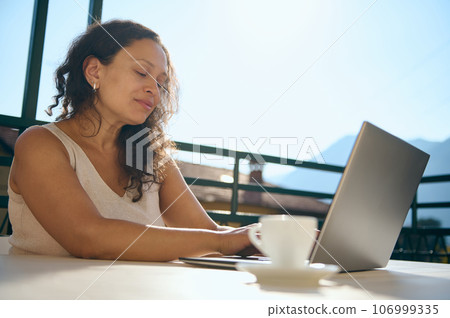 Mature female entrepreneur, businesswoman using laptop computer, working remotely, sitting at table with a coffee cup 106999335