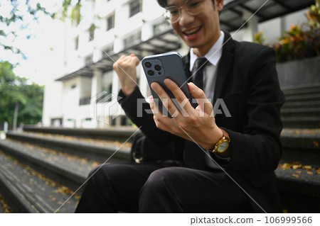 Exited businessman looking at smartphone and celebrating achievement outdoor. Good news, successful, startup 106999566