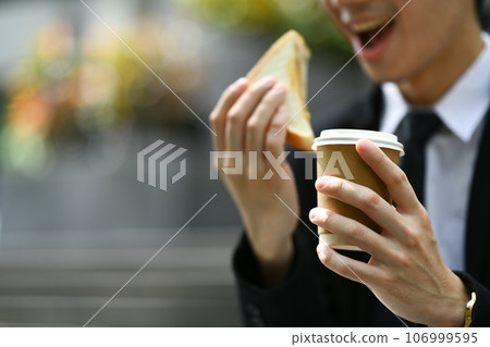 Cropped shot of male office worker in formal wear sitting on stairs outside office and eating tasty sandwiches during lunch 106999595