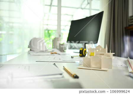 Architect desk with house model, blueprints, safety helmet and computer monitor on white table Architect desk with house model, blueprints, safety helmet and computer monitor on white table 106999624