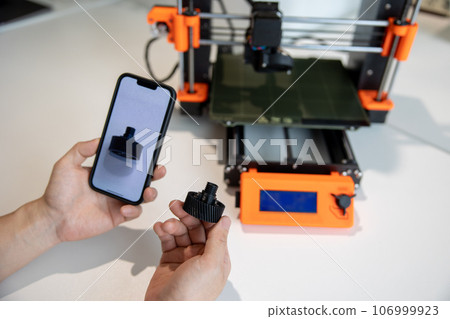 Man holding smartphone with detail on screen, working on 3D printer in creative lab. Man holding smartphone with detail on screen, working on 3D printer in creative lab. 106999923