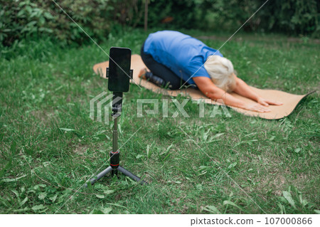 Fit mature 50s female in casual clothes exercising outdoors, practicing yoga on mat and talking at smartphone camera, making video content for followers, looking at webcam on mobile phone on tripod 107000866