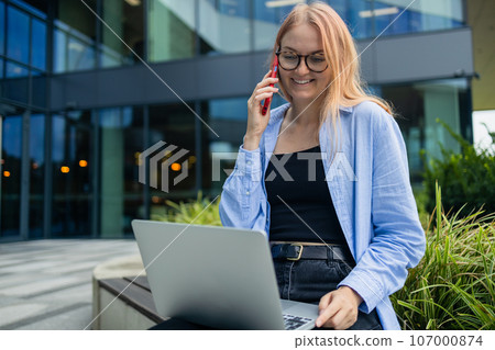 Happy Caucasian student freelancer blonde woman rest use laptop pc computer talk by mobile cell phone look, sitting outdoors against blurred city building. She is keyboarding on laptop. Freelance work Happy Caucasian student freelancer blonde woman rest use laptop pc computer talk by mobile cell phone look, sitting outdoors against blurred city building. She is keyboarding on laptop. Freelance work 107000874