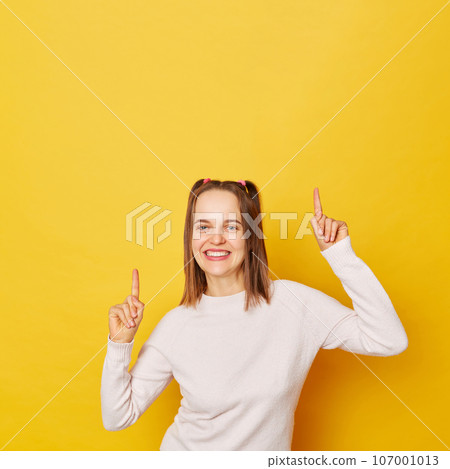 Charming teenage girl in jumper with ponytails isolated over yellow background pointing upwards with broad smile recommending awesome product suggesting click banner showing advertisement promo. Charming teenage girl in jumper with ponytails isolated over yellow background pointing upwards with broad smile recommending awesome product suggesting click banner showing advertisement promo. 107001013
