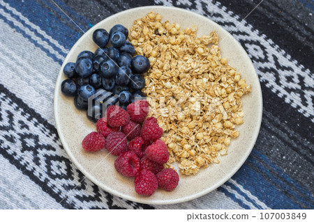 Muesli with blueberries and raspberries on tablecloth background. Granola with wild berries on towel pattern. Healthy eating. Bowl of oat flakes and blueberry and raspberry. Vitamin breakfast. Muesli with blueberries and raspberries on tablecloth background. Granola with wild berries on towel pattern. Healthy eating. Bowl of oat flakes and blueberry and raspberry. Vitamin breakfast. 107003849