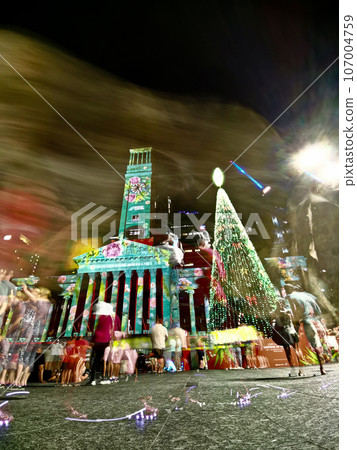 Christmas celebration scene at night at Brisbane City Hall. There is a large Christmas tree with light decoration. There are many movement of people in the photo. 107004759