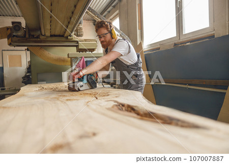 Bearded male wood worker using electric sander for wood. Carpentry workshop. High quality photo 107007887