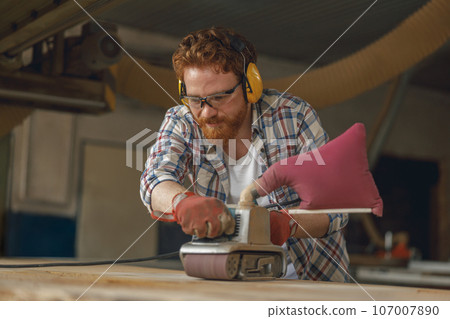 Bearded male wood worker using electric sander for wood. Carpentry workshop. High quality photo 107007890