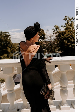 Stylish woman in the city. Fashion photo of a beautiful model in an elegant black dress posing against the backdrop of a building on a city street 107009059