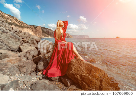 Red dress sea woman. A blonde with flowing hair in a long red dress sits on a rock near the sea. The concept of trips, a photo shoot at the sea Red dress sea woman. A blonde with flowing hair in a long red dress sits on a rock near the sea. The concept of trips, a photo shoot at the sea 107009060