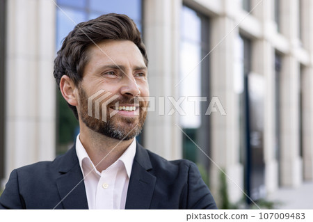 Close-up photo of a young handsome businessman man in a business suit standing outside an office center and looking away with a smile. Close-up photo of a young handsome businessman man in a business suit standing outside an office center and looking away with a smile. 107009483