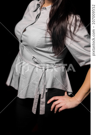 Brunette girl posing with long hair in a light shirt with a pattern of white small polka dots, clothes with push-up breasts on a dark background 107009552