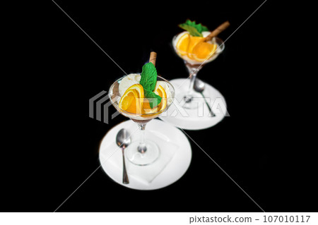 Beautiful serving of scoops of ice cream with slices of orange and mint, ice cream in a glass and saucer with a spoon on a black background 107010117