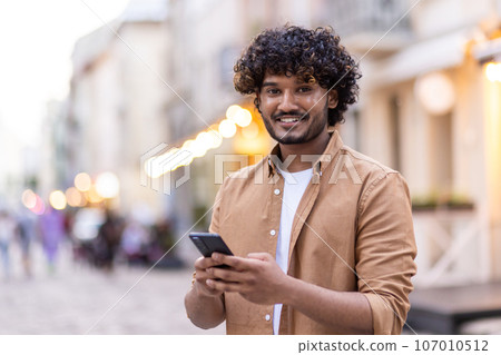 Portrait of a smiling young Indian man traveling tourist standing in the middle of the street, using the phone and smiling at the camera. 107010512