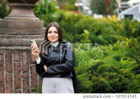 A young pretty girl wearing a black leather jacket walks down the street in a park and uses her smartphone for work. The work of a freelancer and blogger. A young pretty girl wearing a black leather jacket walks down the street in a park and uses her smartphone for work. The work of a freelancer and blogger. 107011545