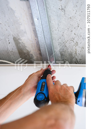Close-up of a worker fastening a suspended ceiling panel with a screwdriver. 107011760