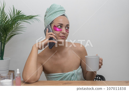 Pretty woman with pink under-eye patches talking by phone and holding cup at table positive lady enjoying skin pampering procedures during home spa day Pretty woman with pink under-eye patches talking by phone and holding cup at table positive lady enjoying skin pampering procedures during home spa day 107011838