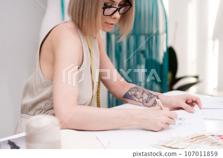 Young blonde woman with tattoo designs glamorous clothes at table in fashion atelier skilled dressmaker works upon new outfit models in sewing studio designer at work Young blonde woman with tattoo designs glamorous clothes at table in fashion atelier skilled dressmaker works upon new outfit models in sewing studio designer at work 107011859
