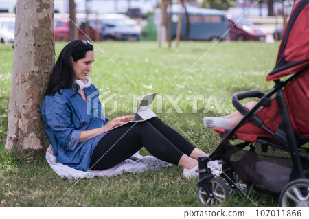 Brunette woman with tablet computer leaning on tree trunk walking with little baby in green city park mother using internet for work while baby sleeping in carriage 107011866