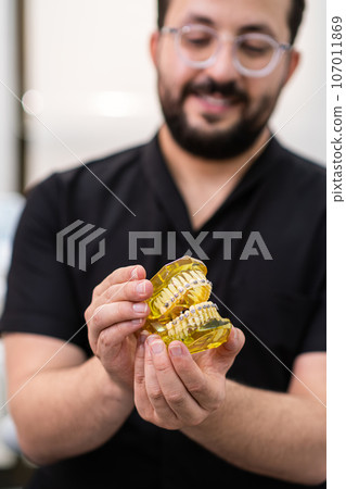 Smiling dentist in black uniform showing open artificial human jaws with installed dental braces for teeth correction bearded doctor consulting patient about orthodontic cases Smiling dentist in black uniform showing open artificial human jaws with installed dental braces for teeth correction bearded doctor consulting patient about orthodontic cases 107011869