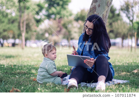 Woman showing gesture calm down to little son talking by phone with colleague and holding tablet sitting in tree shadow in public garden freelancer with dedicated son 107011874