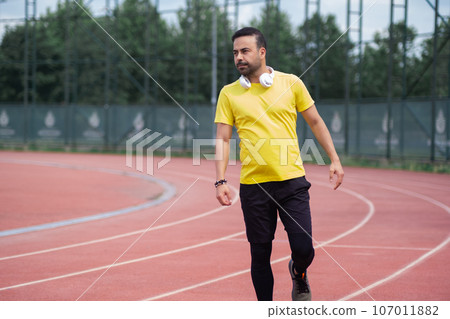 Bearded man in yellow t-shirt with wireless headphones standing on empty running track with red surface and white marking at contemporary sports ground in urban park 107011882