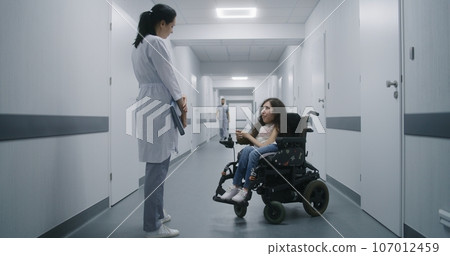 Hospital corridor: Woman with SMA in wheelchair talks to doctor 107012459